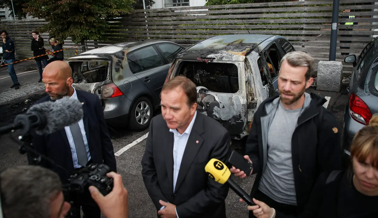 PM Swedia Stefan Lofven memberikan keterangan dengan latar belakang mobil yang hangus dan rusak di kota Gothenburg, Selasa (14/8). Menurut laporan, kelompok gangster menyiram bensin ke atap mobil lalu membakarnya. (Henrik BRUNNSGARD/TT News Agency/AFP)