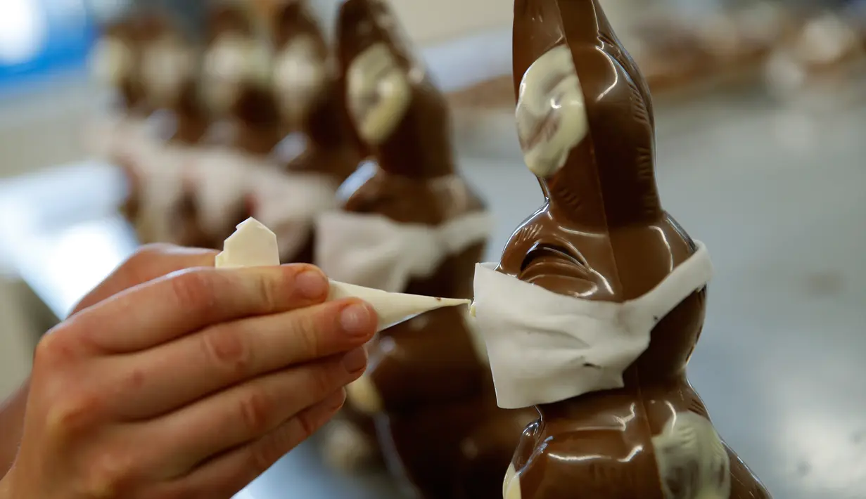 Pekerja menyelesaikan pembuatan cokelat paskah berbentuk kelinci yang mengenakan masker di toko roti Baeckerei Bohnenblust di Bern, Swiss, Jumat (27/3/2020). Di kemasannya dipasang sebuah kertas bertuliskan cara melindungi diri untuk mencegah penularan virus corona. (STEFAN WERMUTH/AFP)