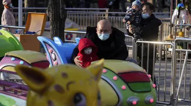 Seorang pria yang mengenakan masker menggendong seorang anak menonton keluarga naik kereta mainan di sebuah taman hiburan di Beijing (17/1/2022). Jumlah bayi yang lahir di China terus menyusut tahun lalu, seiring menyusutnya jumlah tenaga kerja. (AP Photo/Andy Wong)