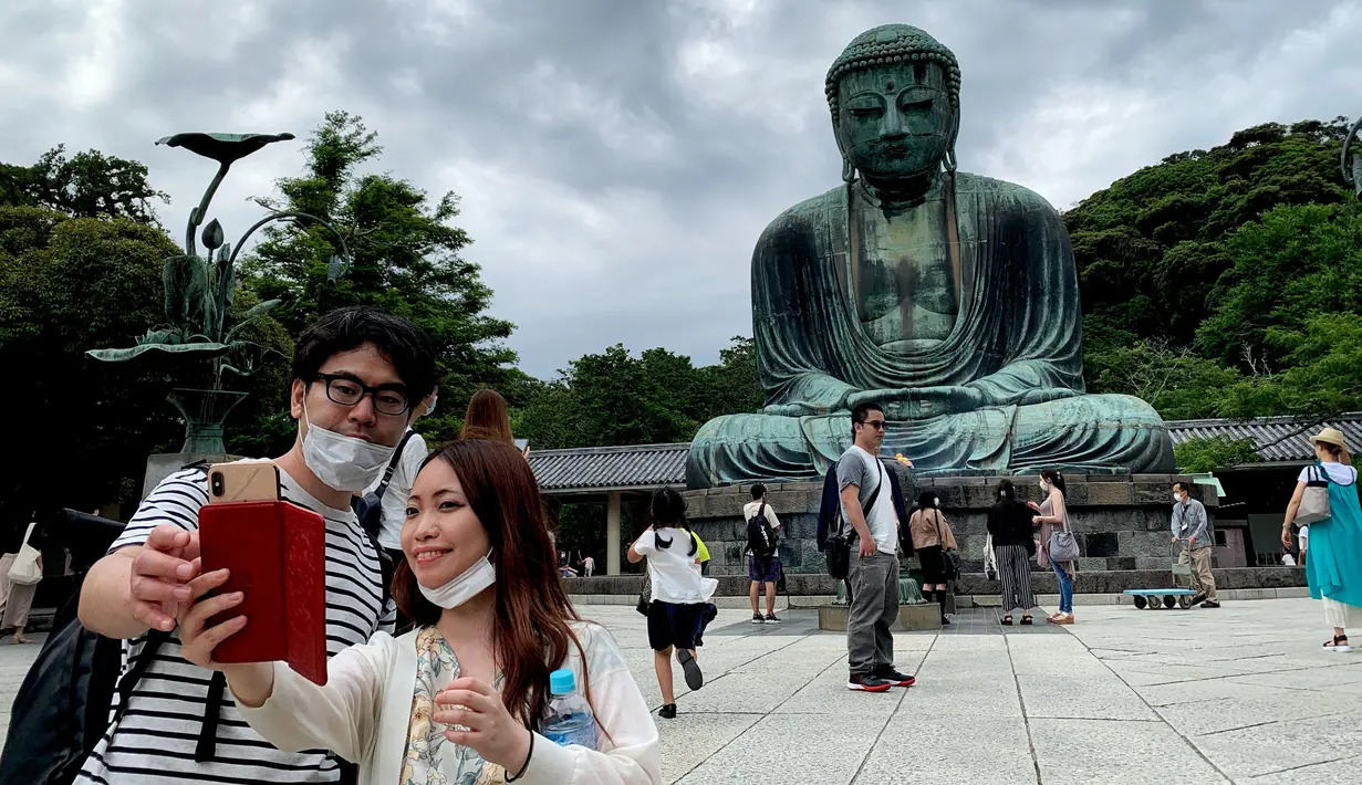 Sepasang pengunjung berswafoto dengan latar belakang The Great Buddha atau yang biasa disebut Kamakura Daibutsu di kuil Kotoku-in di Kamakura, Prefektur Kanagawa, Jepang, Sabtu (20/6/2020). Patung Buddha yang menjulang dengan tinggi 13,35 meter ini didirikan tahun 1252. (Behrouz MEHRI/AFP)