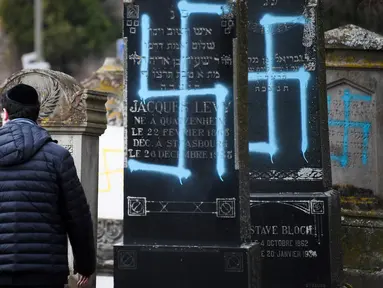 Seorang pria berjalan melewati makam Yahudi yang dicoret simbol Nazi di pemakaman Yahudi, Quatzenheim, Prancis, Selasa (19/2). Aksi vandalisme ini dilakukan terhadap sekitar 80 makam. (Frederick FLORIN/AFP)