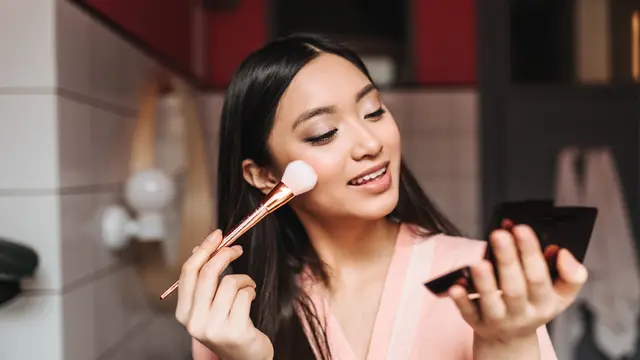 Seorang perempuan mengaplikasikan makeup menggunakan brush sambil bercermin (Foto Dok: Freepik/lookstudio)