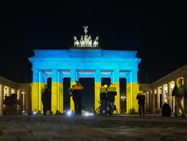 Orang-orang melihat Gerbang Brandenburg yang diterangi dengan warna bendera Ukraina di Berlin, Jerman, Rabu (23/2/2022). Salah satu bangunan ikonik itu diterangi warna bendera nasional Ukraina, biru dan kuning untuk menunjukkan solidaritas selama ketegangan dengan Rusia. (AP Photo/Markus Schreiber)