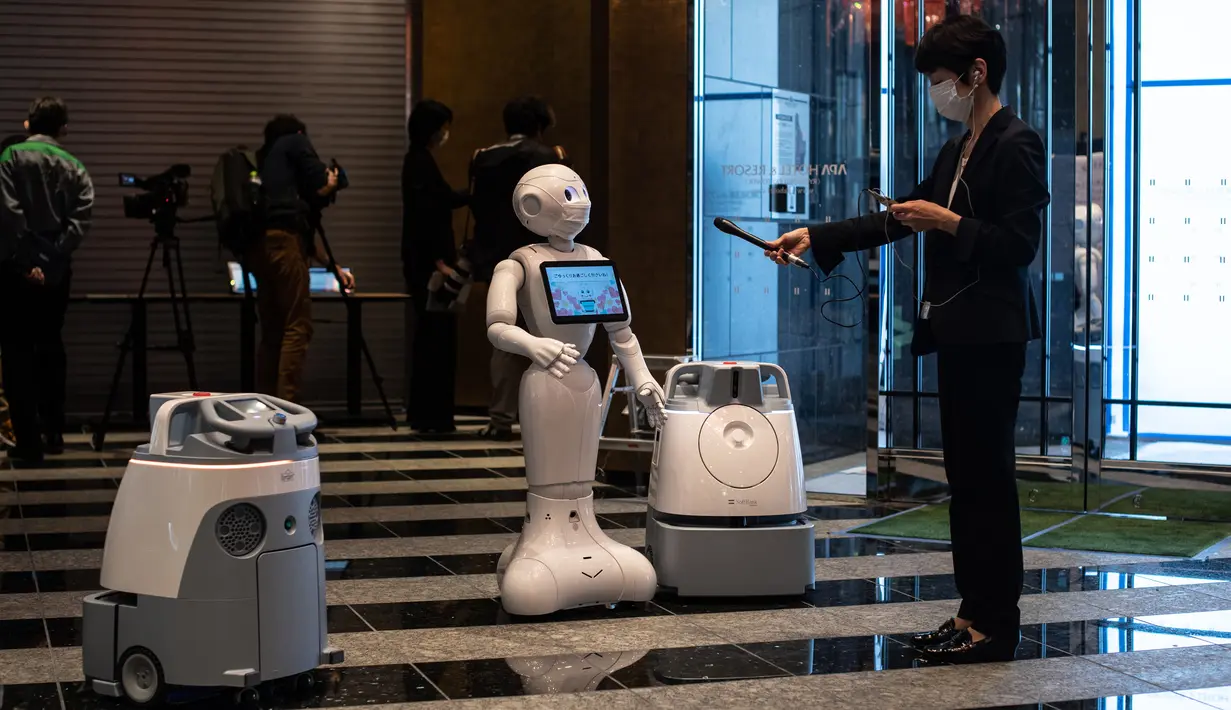 Seorang reporter (kanan) merekam suara salam dari robot Pepper (tengah) dan dua robot pembersih di hotel yang diubah menjadi lokasi karantina pasien COVID-19, Tokyo, Jepang, Jumat (1/5/2020). Sejumlah hotel menyediakan layanan robot ini untuk meringankan beban rumah sakit. (Philip FONG/AFP)