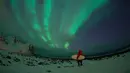 Peselancar berdiri dibawah cahaya Aurora Borealis di pantai bersalju Flackstad di Kepulauan Lofoten, Lingkar Arktik, pada 10 Maret 2016. Peselancar dari seluruh dunia datang ke Lofoten untuk berselancar di kondisi ekstrim. (AFP/Olivier Morin)