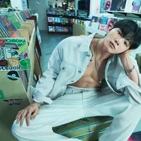 Dalam foto pertama, Jung Kook tampil berani dengan jaket denim putih yang dibiarkan terbuka tanpa inner, memperlihatkan torso atletisnya. Dipadukan dengan jeans senada berpotongan relaxed, gaya ini memancarkan maskulinitas raw yang tetap clean berkat palet monokrom. Pose santai di dalam toko piringan hitam memberi nuansa edgy sekaligus youthful.