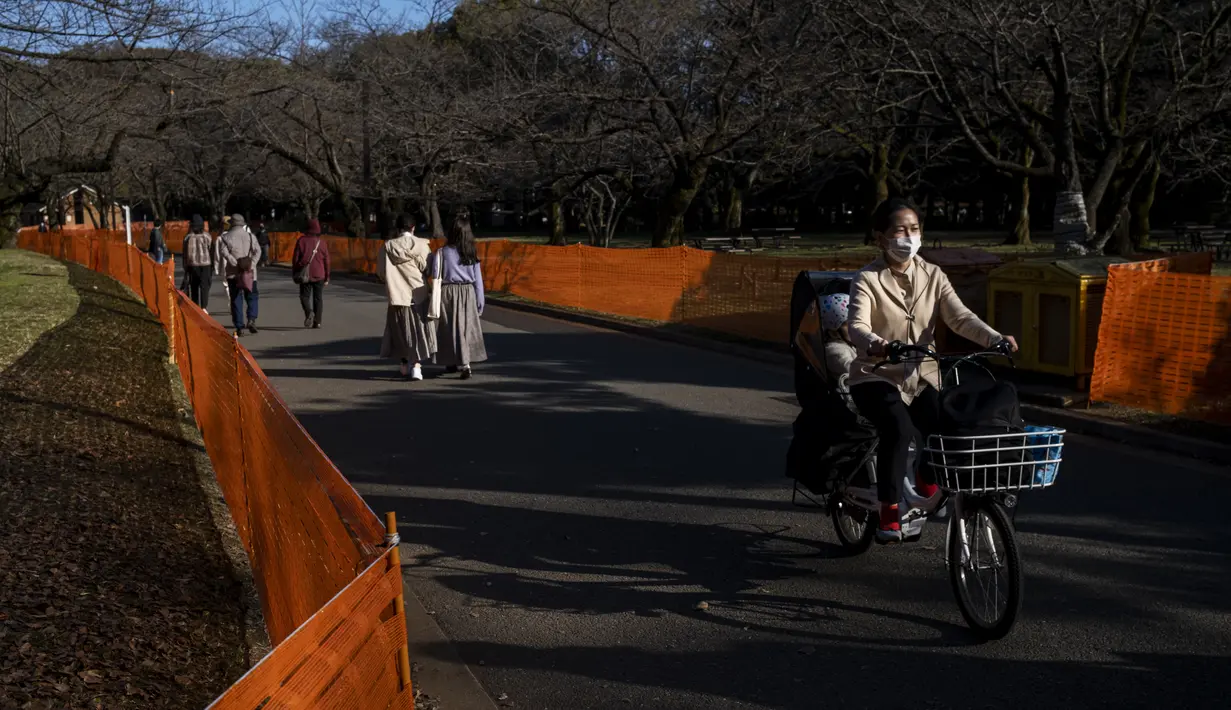 Orang-orang melewati jalan setapak di antara pagar yang ditempatkan untuk menutup bagian taman untuk mencegah pesta melihat bunga sakura (Hanami) di bawah pohon untuk pencegahan keamanan terhadap COVID-19 di salah satu tempat Hanami paling populer di Tokyo (13/3/2021).  (AP Photo/Kiichiro Sato)