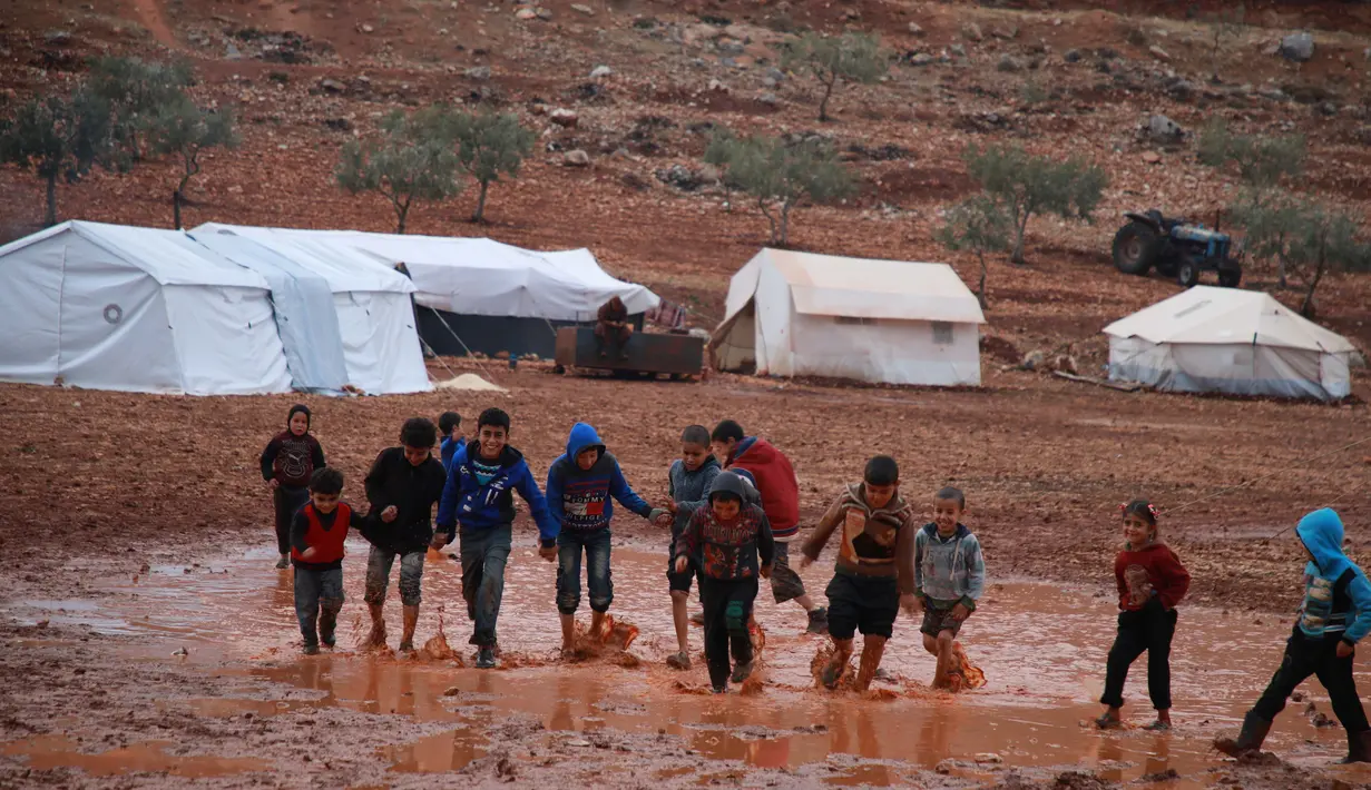 Anak-anak berjalan melewati genangan air hujan di depan tenda di sebuah kamp untuk warga Suriah yang kebanjiran di dekat desa Killi di utara provinsi Idlib barat laut (5/12/2019). (AFP Photo/Aaref Watad)