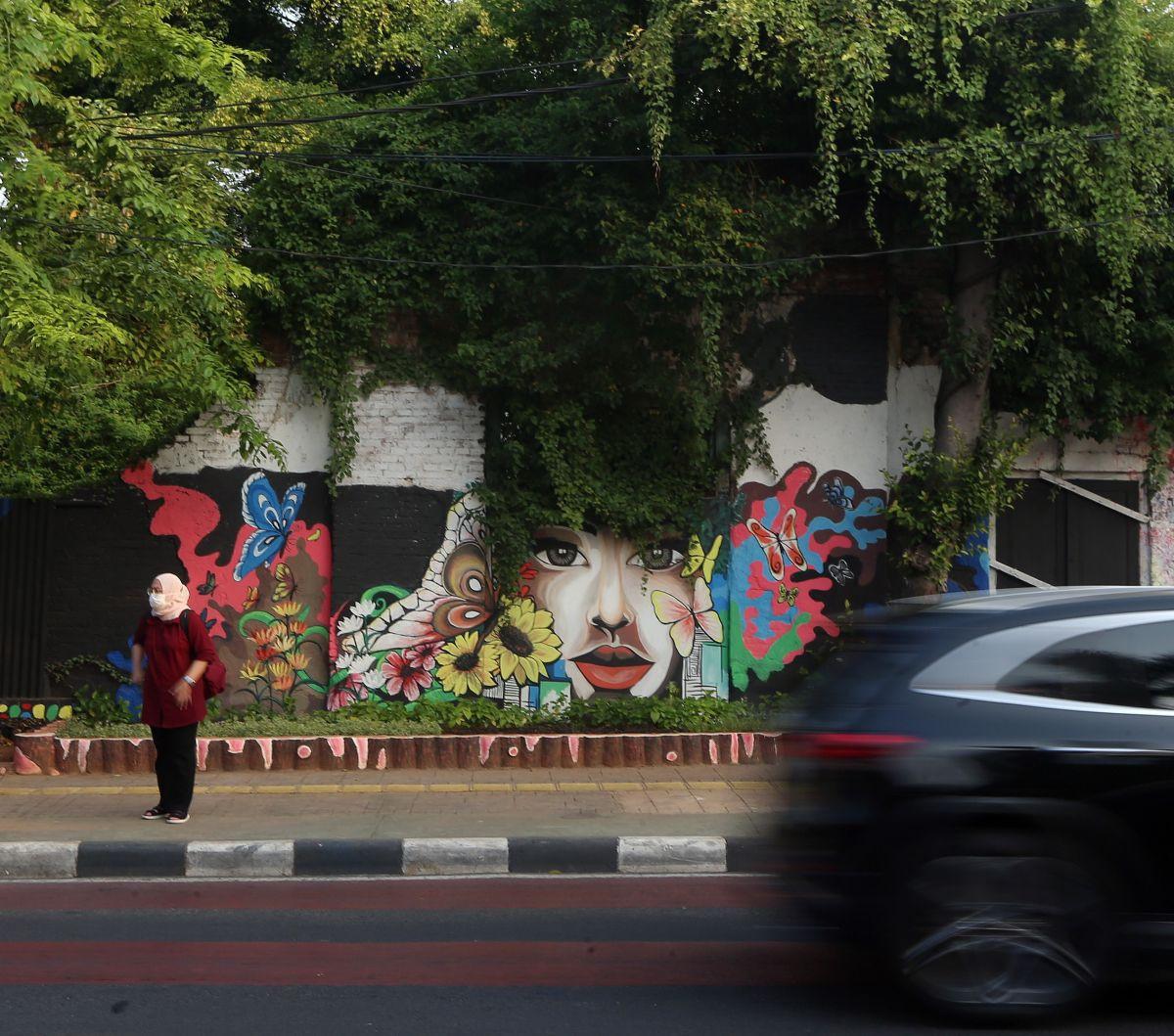 Seorang warga tampak sedang menunggu kendaraan umum di trotoar dengan latar belakang pemandangan mural karya petugas PPSU di kawasan Jalan Malaka, Jakarta.