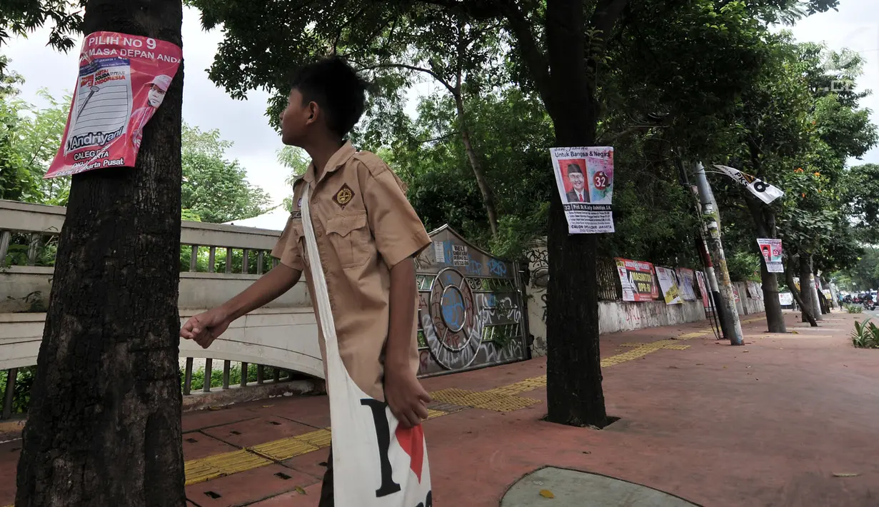 Pelajar melintasi pepohonan yang dipenuhi alat peraga kampanye (APK) calon anggota legislatif (caleg) di Jakarta, Rabu (20/3). Pemasangan APK tersebut melanggar aturan PKPU Nomor 23 Pasal 31 Tentang Bahan Kampanye. (merdeka.com/Iqbal Nugroho)