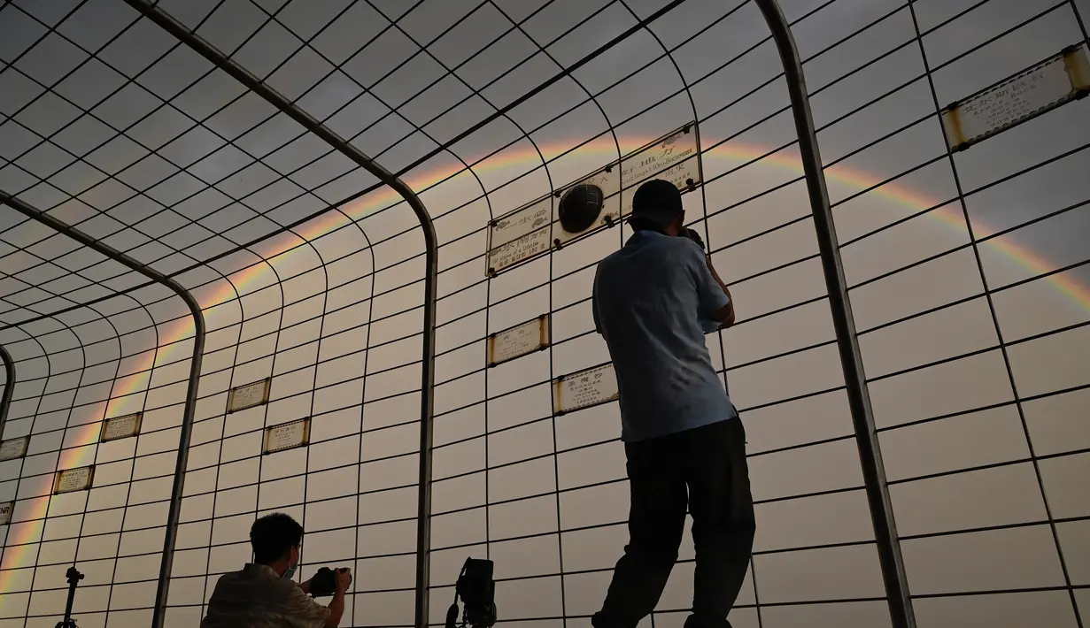 Orang-orang mengambil foto pelangi melalui pagar di Central Television Tower di Beijing (26/8/2021). Keindahan pelangi ganda dimanfaatkan warga Beijing untuk diabadikan lewat foto dan berfoto dengan latar pelangi itu. (AFP/Jade Gao)