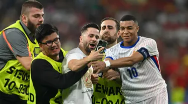 Petugas keamanan menghentikan penyerbu lapangan saat ia mengambil swafoto dengan Kylian Mbappe pada semifinal Euro 2024 antara Spanyol vs Prancis di Allianz Arena, Rabu (10/7/2024) dini hari WIB. (Fabrice COFFRINI / AFP)