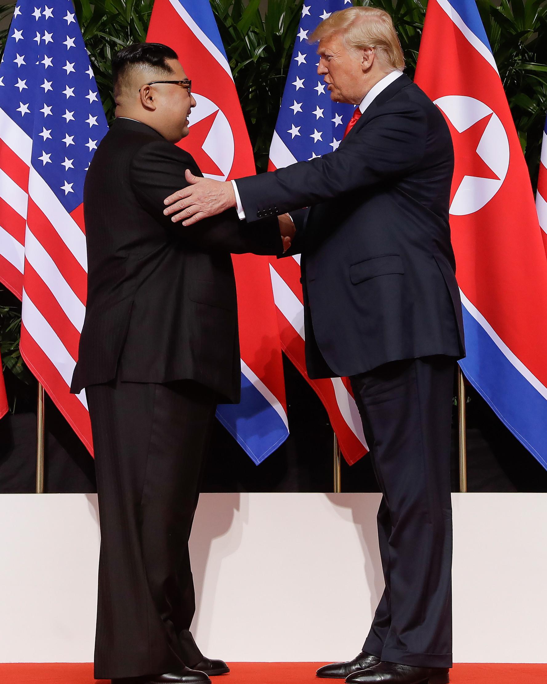 Presiden AS Donald Trump  berjabat tangan dengan Pemimpin Korea Utara, Kim Jong-un dalam pertemuan bersejarah di resor Capella, Pulau Sentosa, Selasa (12/6). Kim dan Trump hadir di depan jurnalis dengan latar belakang bendera Korut dan AS. (AP/Evan Vucci)