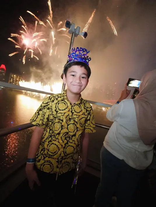 Rafathar tebar senyum tipisnya sambil kenakan kemeja Barocco Kids Shirt Versace seharga Rp5,8 jutaan. Kemeja dengan motif print itu dipadu dengan celana pendek dan aksesori tahun baru [@raffinagita1717]