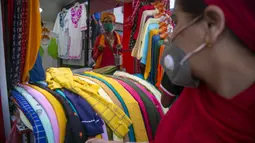 Seorang wanita yang mengenakan masker sebagai tindakan pencegahan terhadap virus corona COVID-19 memeriksa barang-barang yang dipajang di toko pakaian saat seorang lelaki suci menunggu sedekah di pintu masuknya, Kathmandu, Nepal, Selasa (13/10/2020). (AP Photo/Niranjan Shrestha)