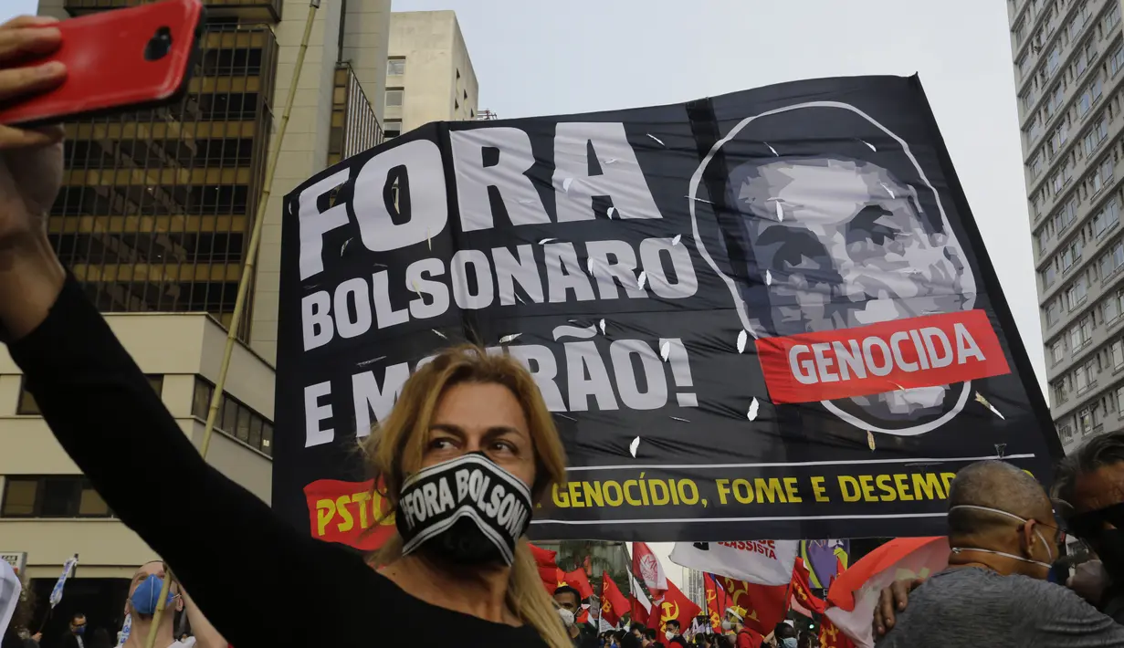 Seorang perempuan berswafoto dengan latar belakang spanduk yang menggambarkan Presiden Brasil Jair Bolsonaro dan sebuah pesan dalam bahasa Portugis, "Keluar Bolsonaro!" selama aksi protes menuntut Bolsonaro mundur, di Sao Paulo, Sabtu (3/7/2021). (AP Photo/Nelson Antoine)