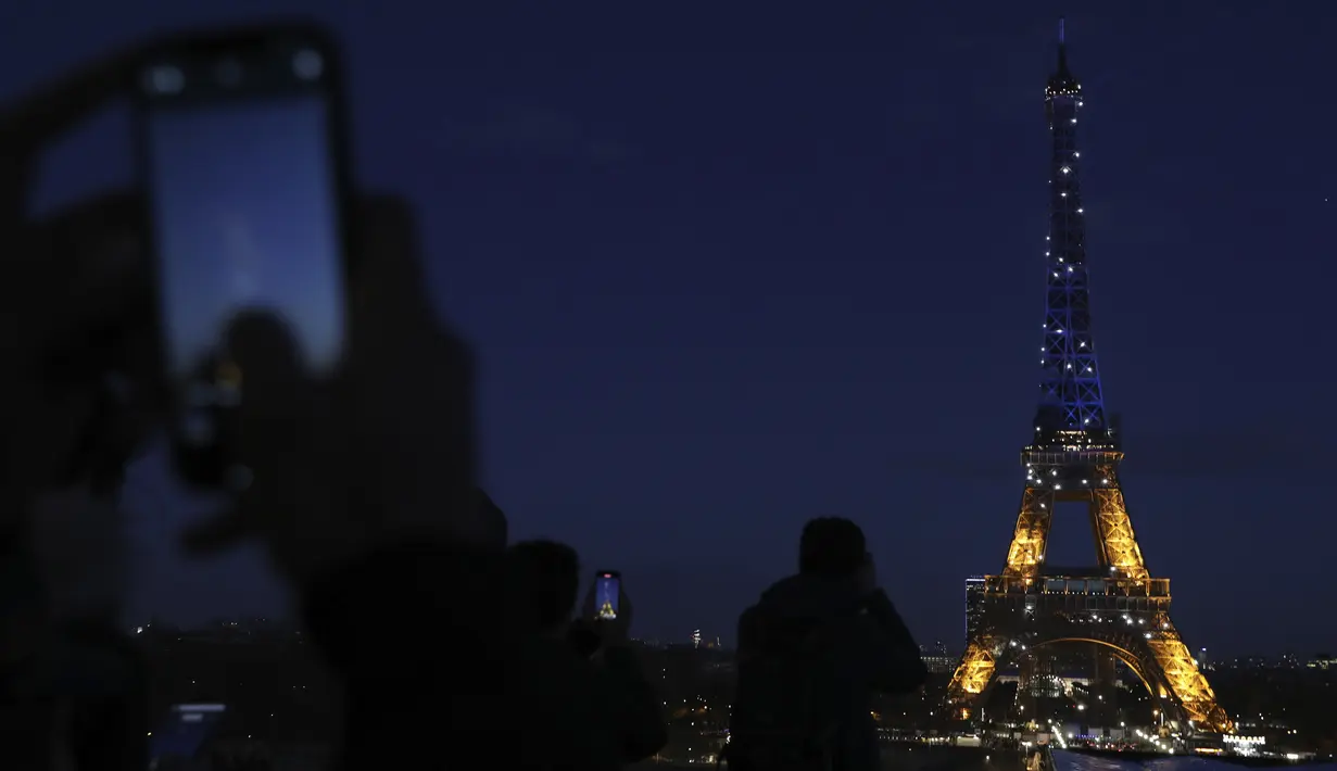 Warga memotret Menara Eiffel yang diterangi warna bendera Ukraina di Paris (25/2/2022). Pasukan Rusia menyerang ibu kota Ukraina pada Jumat (25/2) dengan tembakan dan ledakan dalam invasi ke negara demokratis yang memicu kekhawatiran perang yang lebih luas di Eropa. (AP Photo/Adrienne Surprenant)