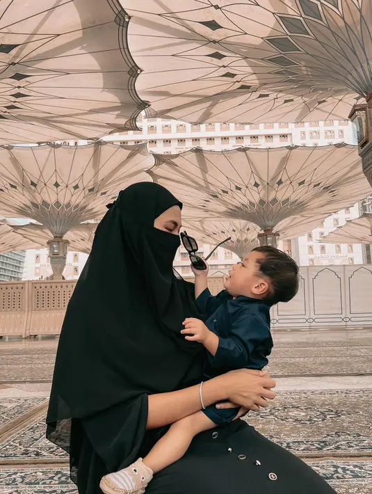 Potret gemas, Paula Verhoeven yang menjalankan ibadah Umroh membawa sang buah hati. Di foto ini, ia menggendong anak keduanya, mengenakan gamis hitam polos dan hijab bercadar yang serasi. [Foto: Instagram/paula_verhoeven]