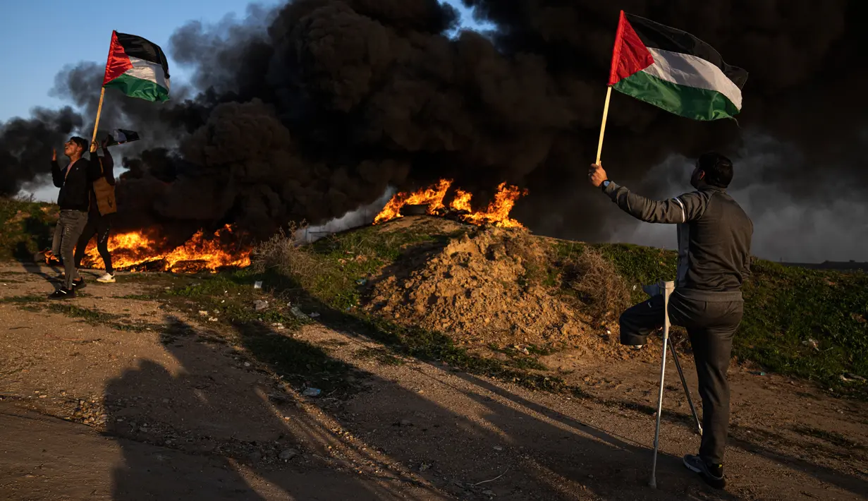 Warga Palestina membakar ban dan mengibarkan bendera nasional saat protes terhadap serangan militer Israel terhadap kamp pengungsi Jenin di perbatasan Israel-Palestina, sebelah timur Kota Gaza, 26 Januari 2023. Serangan Israel terhadap kamp pengungsi Jenin menjadi salah satu pertempuran paling mematikan dalam beberapa tahun terakhir. (AP Photo/Fatima Shbair)