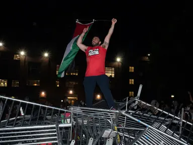 Seorang mahasiswa Universitas George Washington melambaikan bendera Palestina saat berdiri di atas barikade polisi yang diterobos untuk menduduki University Yard dalam aksi protes pro-Palestina di Washington, Minggu, 28 April 2024. (AP Photo/Cliff Owen)