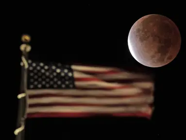 Bayangan bumi menutupi bulan purnama selama gerhana bulan sebagian atau parsial saat terbenam melewati bendera AS di atas sebuah gedung di pusat kota Kansas City, Missouri, Jumat (19/11/2021). (AP Photo/Charlie Riedel)