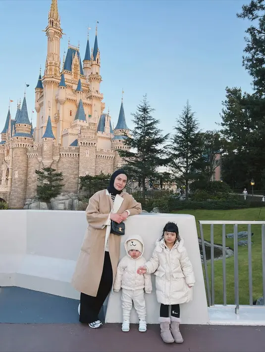 Di depan istana Disneyland, Dwi Handayani berpose bersama 2 buah hatinya. Dwi Handayani mengenakan long coat cokelat yang dipadu dengan celana panjang hitam. Sedangkan kedua anaknya terlihat mengenakan outfit senada berwarna putih. Foto: Instagram.