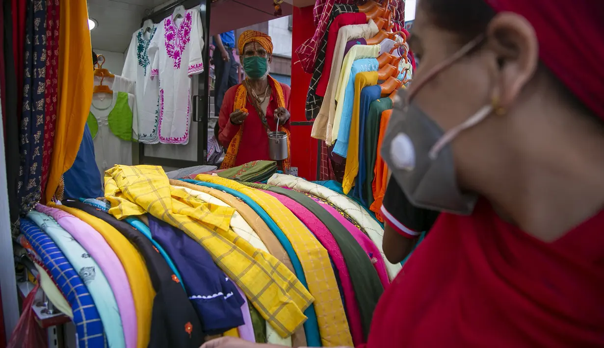 Seorang wanita yang mengenakan masker sebagai tindakan pencegahan terhadap virus corona COVID-19 memeriksa barang-barang yang dipajang di toko pakaian saat seorang lelaki suci menunggu sedekah di pintu masuknya, Kathmandu, Nepal, Selasa (13/10/2020). (AP Photo/Niranjan Shrestha)