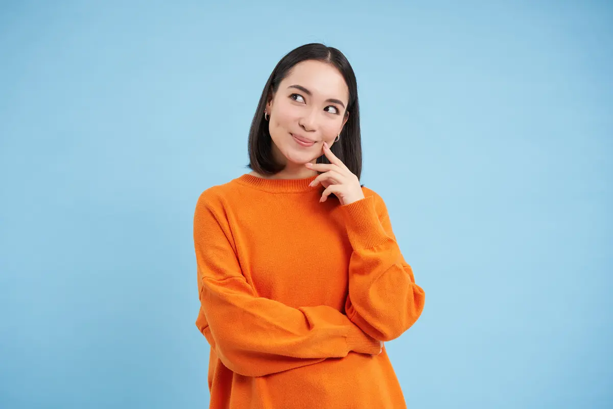 Seorang perempuan berbaju oranye sedang berpikir sambil memegang pipinya menggunakan telunjuk (Foto Dok: Freepik/benzoix)