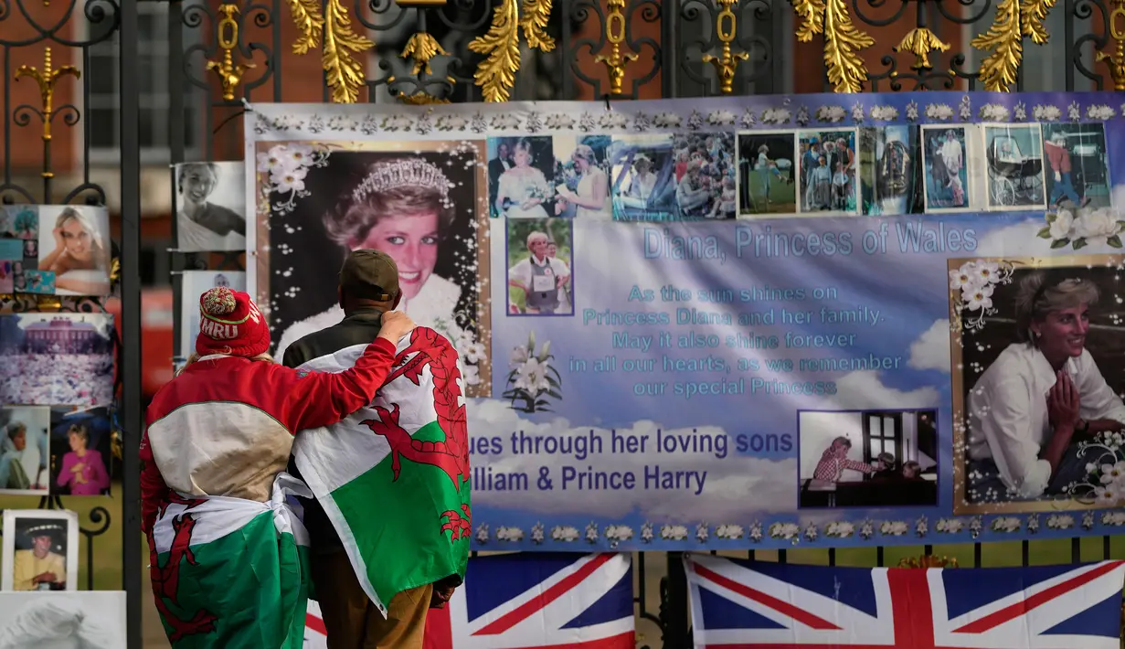 Sepasang kekasih, terbungkus bendera Welsh, melihat potret dan pesan peringatan untuk Putri Diana yang dipajang di gerbang Istana Kensington, London, Rabu (31/8/2022). Hari ini menandai peringatan 25 tahun kematian Putri Diana dalam kecelakaan mobil di Paris. (AP Photo/Alastair Grant)