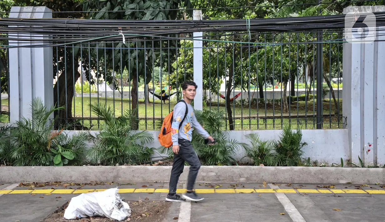 Pejalan kaki melintas di dekat kabel utilitas yang menjuntai di Jalan Proklamasi, Pegangsaan, Kecamatan Menteng, Jakarta Pusat, Kamis (6/10/2022). Minimnya pengawasan dalam tahap penyelesaian proyek revitalisasi trotoar oleh pihak terkait menyebabkan jaringan kabel utilitas di kawasan tersebut semrawut  yang dapat membahayakan keselamatan pengguna jalan, terutama saat musim penghujan. (merdeka.com/Iqbal S. Nugroho)