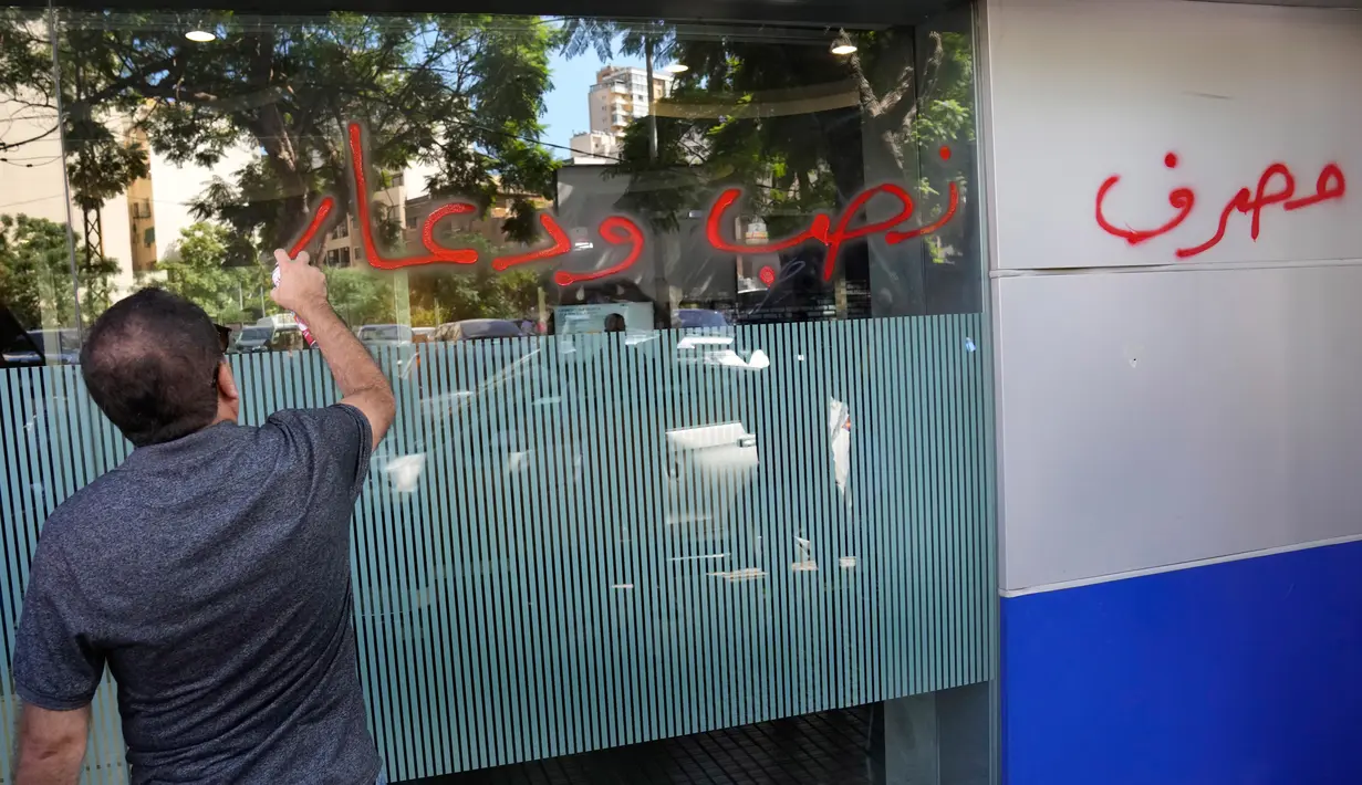 Seorang pria menyemprotkan cairan berwarna merah dan menuliskan kalimat berbahasa Arab yang artinya "Pencurian bank dan prostitusi," di jendela bank yang diserang di Beirut, Lebanon, Rabu (14/9/2022). Akibat kasus ini, muncul kasus pembobolan lain yang terjadi di bank-bank yang ada di Lebanon. (AP Photo/Hussein Malla)