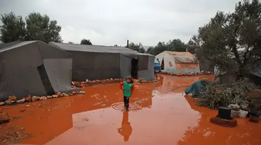 Seorang gadis berjalan melewati genangan air hujan di depan tenda di kamp untuk warga Suriah yang kebanjiran di dekat desa Killi di utara provinsi barat laut Idlib (5/12/2019). (AFP Photo/Aaref Watad)