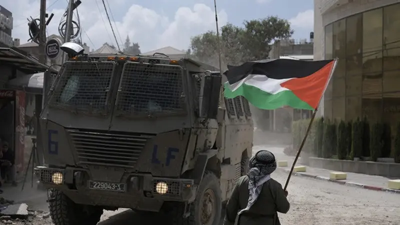 Seorang pria melambaikan bendera Palestina saat kendaraan lapis baja Israel bergerak selama operasi militer di kamp pengungsi Nur Shams, Tulkarem, Tepi Barat yang diduduki pada Kamis (29/8/2024).