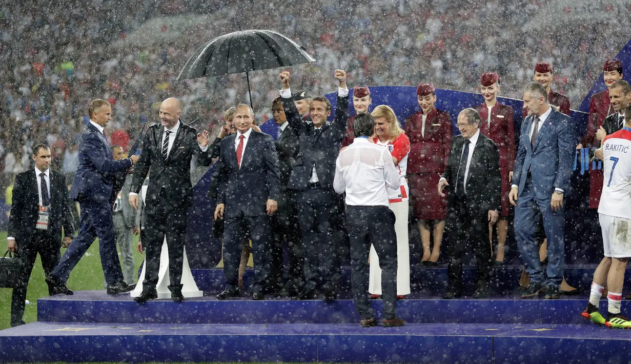 Presiden Rusia, Vladimir Putin dilindungi oleh seorang pemandu yang memegang payung sementara para pejabat lainnya basah kuyup saat hujan turun selama penyerahan medali dan trofi Piala Dunia 2018 di Stadion Luzhniki, Minggu (15/7). (AP/Matthias Schrader)