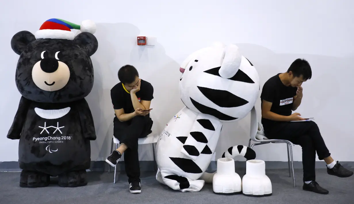 Para pekerja melihat ponsel mereka di samping maskot Olimpiade Musim Dingin dan Paralimpiade Pyeongchang 2018 di dekat stan Korea Selatan selama Pameran Olahraga Musim Dingin Dunia di Beijing, 7 September 2017. (AP Photo/Andy Wong)