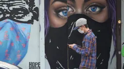 Seorang pria yang mengenakan masker melewati mural yang dilukis pada bisnis papan di pusat kota Austin, Texas, Selasa (9/3/2021). Mandat Gubernur Texas Greg Abbott yang mencabut keharusan mengenakan masker di negara bagiannya akan berlaku penuh mulai 10 Maret. (AP Photo/Eric Gay)