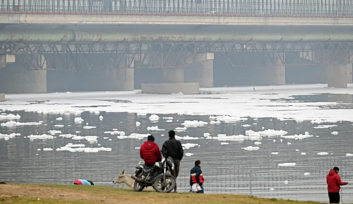 Orang-orang berdiri di tepi Sungai Yamuna yang dilapisi busa tercemar, New Delhi, India, 26 Desember 2021. Menurut para ahli, busa yang mencemari Sungai Yamuna dapat menyebabkan masalah pernapasan dan kulit.(Sajjad HUSSAIN/AFP)
