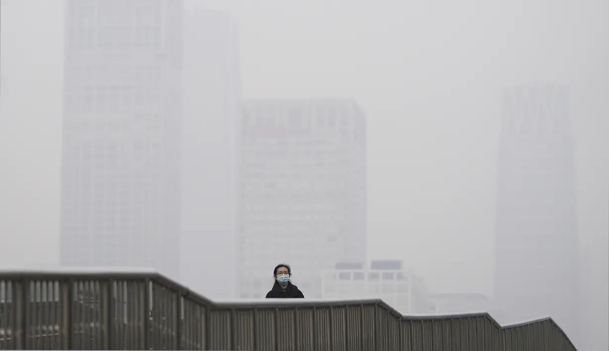 Seorang wanita mengenakan masker untuk membantu mengekang penyebaran virus corona berjalan di jembatan di atas gedung perkantoran di Kawasan Pusat Bisnis yang diselimuti kabut polusi di Beijing, Kamis (18/11/2021).  (AP Photo/Andy Wong)