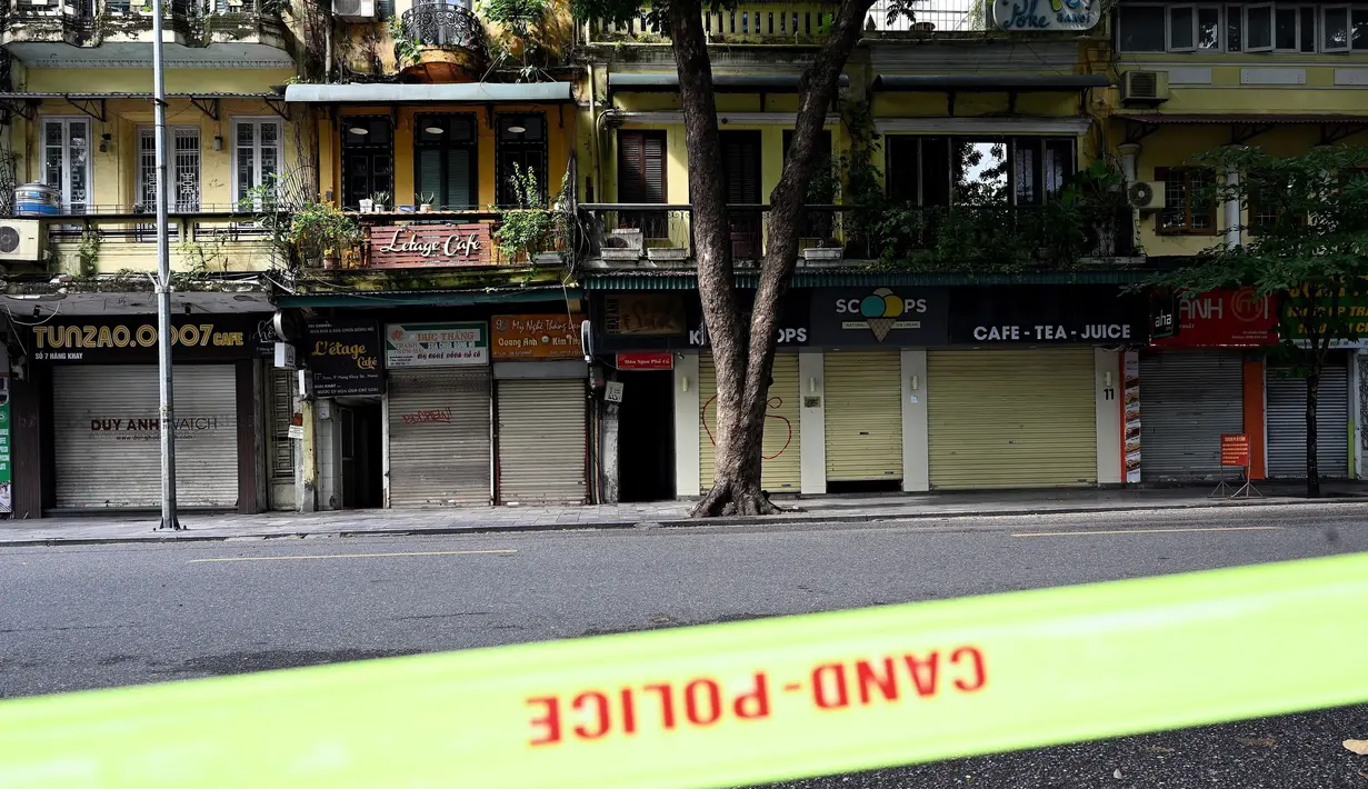 Toko-toko dan restoran-restoran yang tutup terlihat melewati garis polisi di sepanjang jalan yang sepi di Hanoi pada hari pertama lockdown, Sabtu (24/7/2021). Vietnam memberlakukan pembatasan yang lebih ketat di Hanoi akibat gelombang infeksi COVID-19 terburuknya. (Manan VATSYAYANA / AFP)