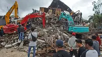 Sejumlah warga menyaksikan alat berat mencari korban di reruntuhan gedung pemerintah daerah yang runtuh saat gempa bumi di Mamuju, Sulawesi Barat, Jumat (15/1/2021). (AP Photo/Azhari Surahman)