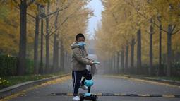 Seorang anak bermasker mengendarai skuter di sepanjang jalan dengan pohon ginkgo pada hari yang berkabut dan tercemar di Beijing pada Sabtu (6/11/2021). Kabut asap tebal menyelimuti sebagian besar China bagian utara, termasuk Beijing. (JADE GAO/AFP)