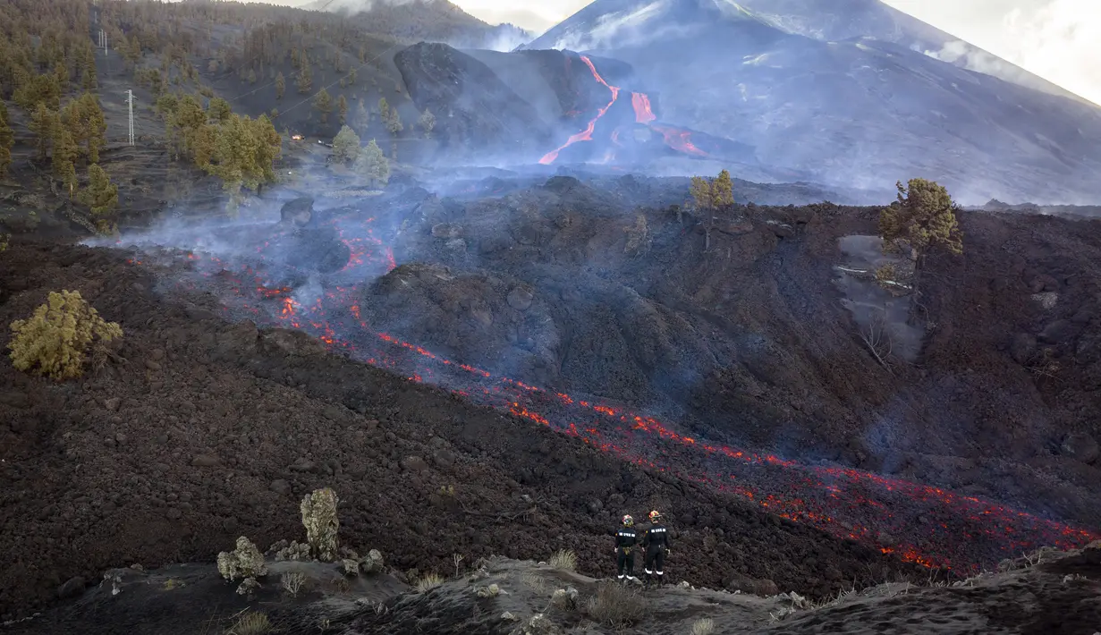 Petugas pemadam kebakaran melihat lava yang mengalir dari gunung berapi di pulau Canary La Palma, Spanyol (30/11/2021). Beberapa lubang vulkanik baru dibuka di La Palma, melepaskan lava baru yang mengalir cepat menuruni punggung bukit. (AP/Emilio Morenatti)