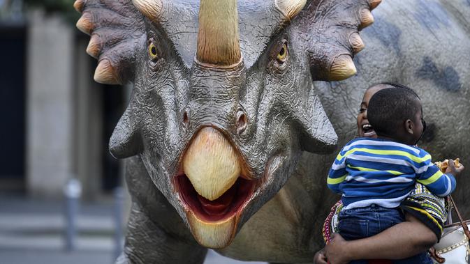 Seorang wanita mengendong anaknya berjalan di samping replika dinosaurus Styracosaurus seukuran aslinya di pusat kota Bochum, Jerman (12/8/2019). Pameran ini menampilkan 33 replika dinosaurus di sekitar pusat kota, berada di Bochum. (AP Photo/Martin Meissner)