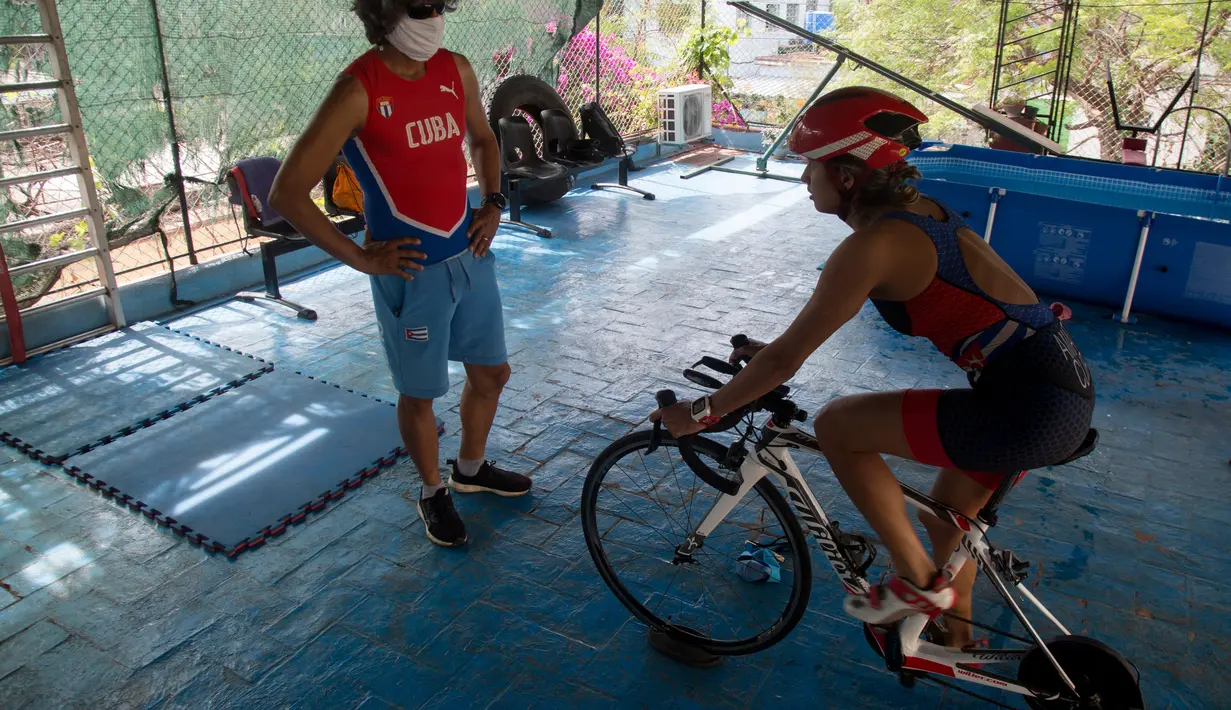 Atlet triathlon Kuba, Leslie Amat mengendarai sepedanya, diadaptasi menjadi stasioner, di bawah pengawasan pelatihnya Dioseles Fernandez di teras rumahnya di Havana, Senin (20/4/2020). Atlet Kuba tak bisa berlatih di fasilitas olahraga seperti biasanya selama pademi corona. (AP /Ismael Francisco)