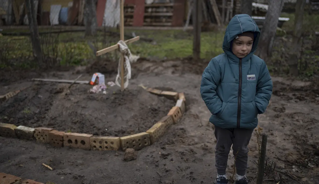 Di halaman rumahnya, Vlad Tanyuk (6) berdiri di dekat makam ibunya Ira Tanyuk, yang meninggal karena kelaparan dan stres akibat perang, di pinggiran Kiv, Ukraina, Senin, 4 April 2022. Gundukan tanah yang ditutupi oleh salib kayu sederhana itu menandai makam ibunya. (AP Photo/Rodrigo Abd)