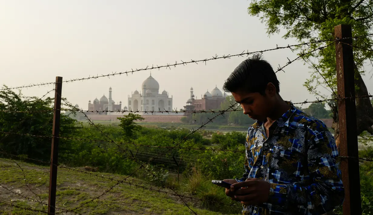 Seorang pria memainkan ponselnya di seberang Taj Mahal dari tepi sungai Yamuna di Agra, Jumat (4/5). Mahkamah Agung India meminta pemerintah mencari cara untuk mengembalikan warna asli situs bersejarah dunia, Taj Mahal. (AFP/CHANDAN KHANNA)