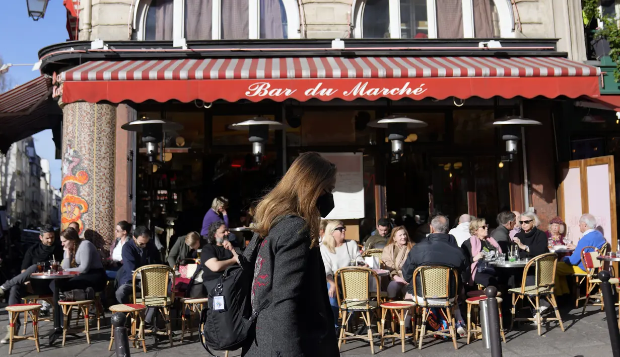 Seorang perempuan mengenakan masker, berjalan di depan kafe Paris yang terbuka, di Paris, Senin (14/3/2022). Prancis telah mencabut sebagian besar pembatasan COVID-19 pada hari Senin, memungkinkan orang melepas masker di hampir semua tempat. (AP Photo/Francois Mori)