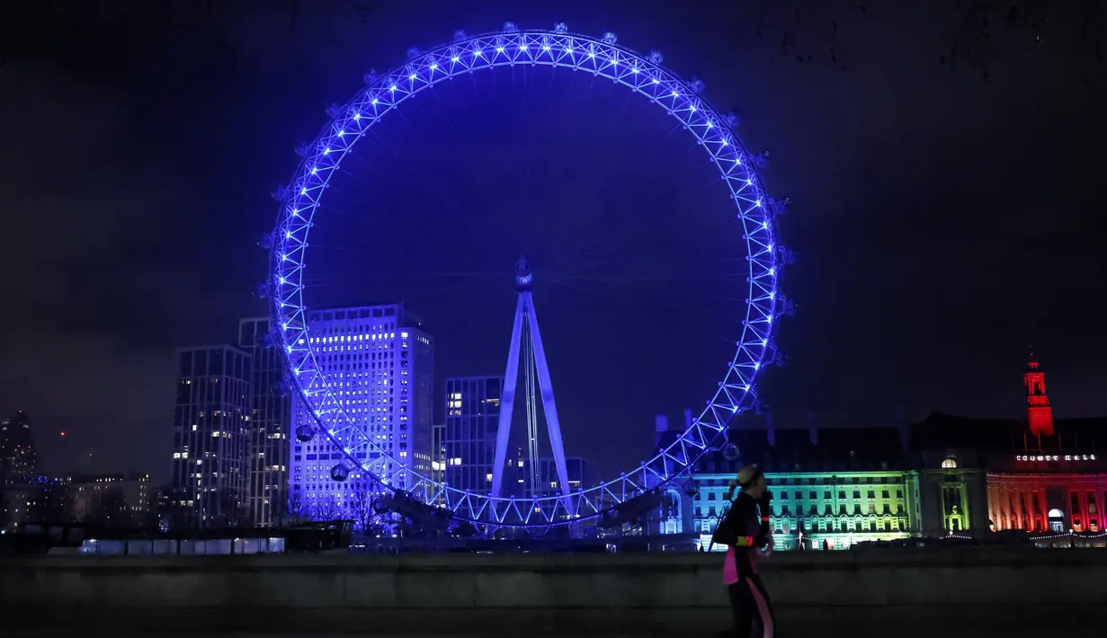 Pejalan kaki melintas di depan London Eye yang diterangi dengan cahaay biru untuk berterima kasih kepada National Health Service (NHS) di London, Kamis (7/1/2021). NHS merupakan sistem layanan kesehatan nasional yang menjadi ujung tombak Inggris dalam memerangi pandemi Covid-19. (Tolga Akmen / AFP)