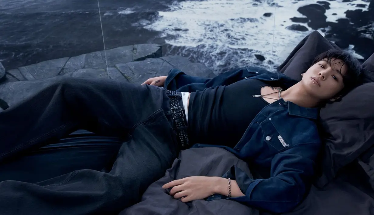 Look berikutnya menghadirkan nuansa misterius lewat outfit serba gelap: tank top hitam, jaket denim biru tua, dan jeans hitam. Pose bersandar dengan mata terpejam menciptakan kesan sensual yang lebih emosional, memperlihatkan sisi artistik Jung Kook. [Dok/Calvin Klein].