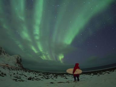 Seorang peselancar berdiri menatap sinar aurora sebelum beraksi di pantai Unstad, Lofoten, Arctic Circle, (10/3/2016). (AFP/Olivier Morin)
